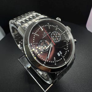 May include: A silver-toned wristwatch with a dark brown face and a silver band. The watch features multiple sub-dials, white dot hour markers, and a date display. The watch is displayed on a clear stand against a black background.
