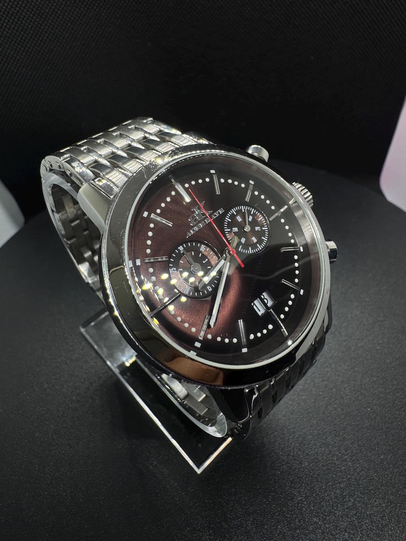 May include: A silver-toned wristwatch with a dark brown face and a silver band. The watch features multiple sub-dials, white dot hour markers, and a date display. The watch is displayed on a clear stand against a black background.
