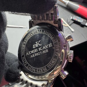 May include: Close-up of the back of a silver stainless steel watch. The watch face is black with the brand name "ADEE KAYE" and model number "AK8027-MB". The watch band is a woven silver design. The watch is water resistant.
