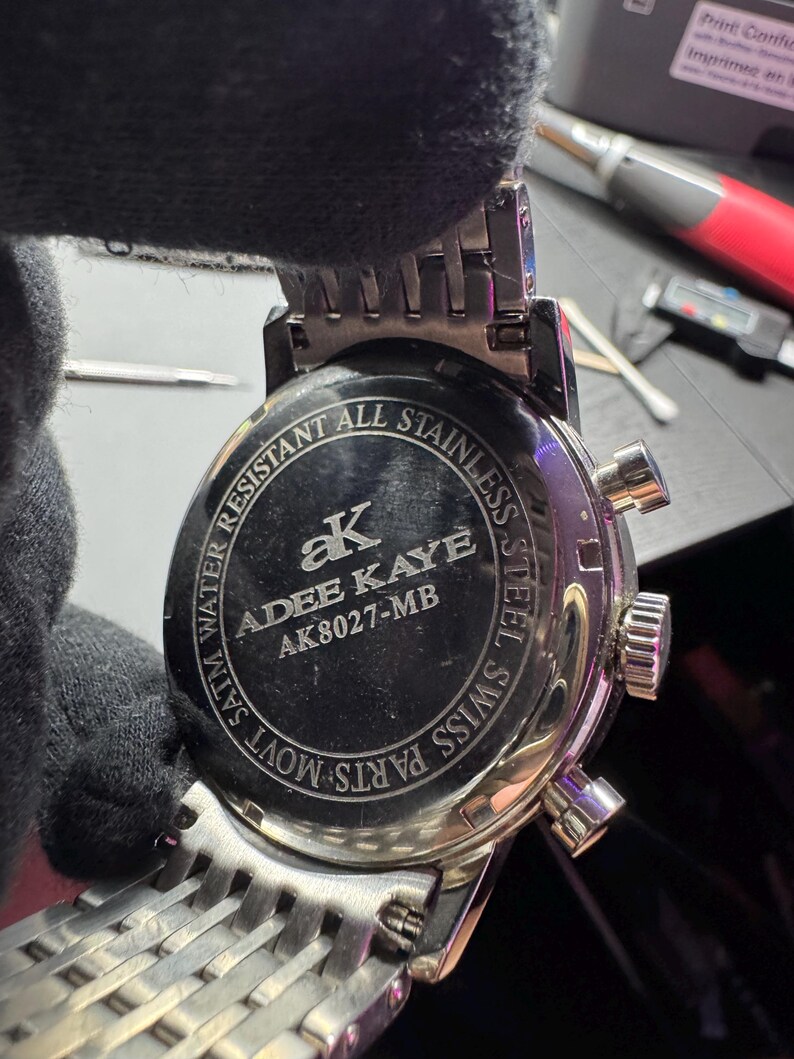 May include: Close-up of the back of a silver stainless steel watch. The watch face is black with the brand name "ADEE KAYE" and model number "AK8027-MB". The watch band is a woven silver design. The watch is water resistant.