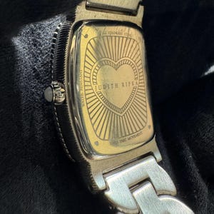 May include: Close-up of a silver and gold-toned watch. The watch back features a heart design with the words "JUDITH RIPKA" and "SWISS PART MOVEMENT". The watch band is silver and the watch is water resistant.