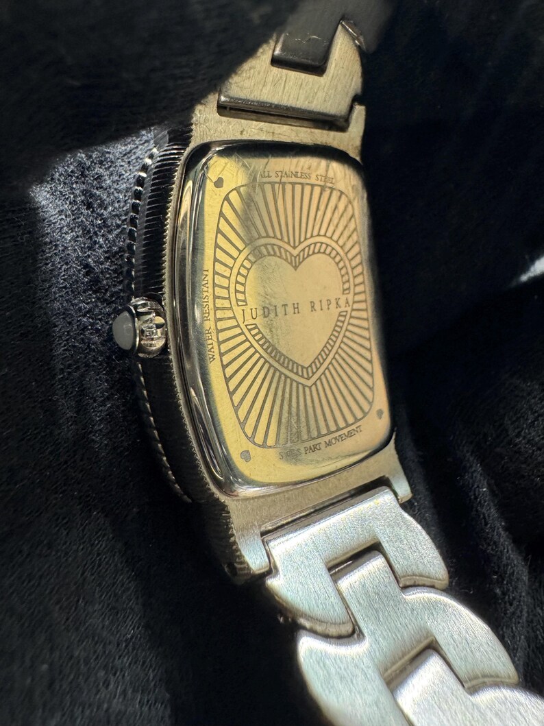 May include: Close-up of a silver and gold-toned watch. The watch back features a heart design with the words "JUDITH RIPKA" and "SWISS PART MOVEMENT". The watch band is silver and the watch is water resistant.