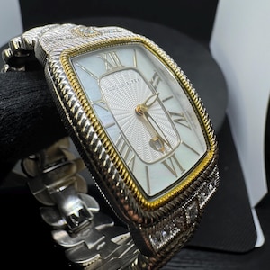 May include: A rectangular wristwatch with a silver and gold-tone case and a white face. The watch features Roman numeral hour markers and a textured centre. The band is silver-toned and has a unique, articulated design. The watch is adorned with small, clear gemstones.