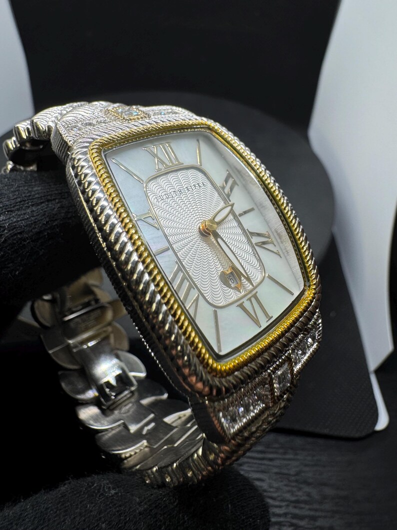 May include: A rectangular wristwatch with a silver and gold-tone case and a white face. The watch features Roman numeral hour markers and a textured centre. The band is silver-toned and has a unique, articulated design. The watch is adorned with small, clear gemstones.