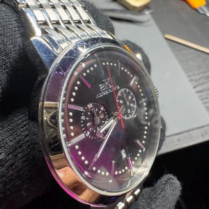 May include: A stainless steel wristwatch with a black face and silver band. The watch features a chronograph design with multiple sub-dials and a red second hand. The watch has a polished bezel and a link-style bracelet.