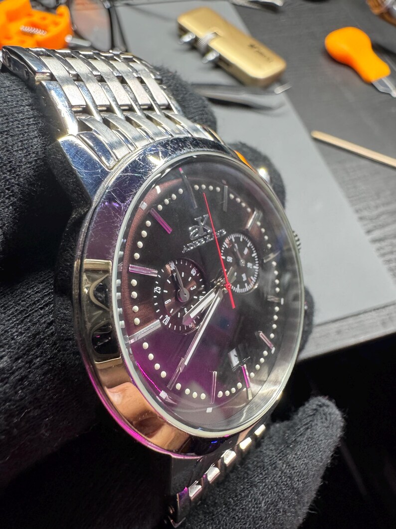May include: A stainless steel wristwatch with a black face and silver band. The watch features a chronograph design with multiple sub-dials and a red second hand. The watch has a polished bezel and a link-style bracelet.