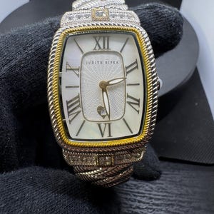 May include: A silver and gold-toned wristwatch with a rectangular face. The watch features Roman numerals, a date display, and the brand name "JUDITH RIPKA". The watchband has a braided design and is adorned with small, sparkling stones.