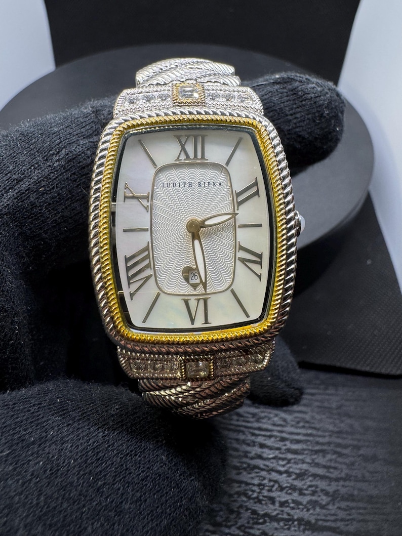 May include: A silver and gold-toned wristwatch with a rectangular face. The watch features Roman numerals, a date display, and the brand name "JUDITH RIPKA". The watchband has a braided design and is adorned with small, sparkling stones.