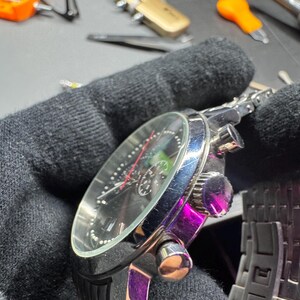 May include: A close-up of a silver and black wristwatch with a black face, featuring silver hands and markers. The watch has a stainless steel band and multiple dials. The watch is held in a black gloved hand.