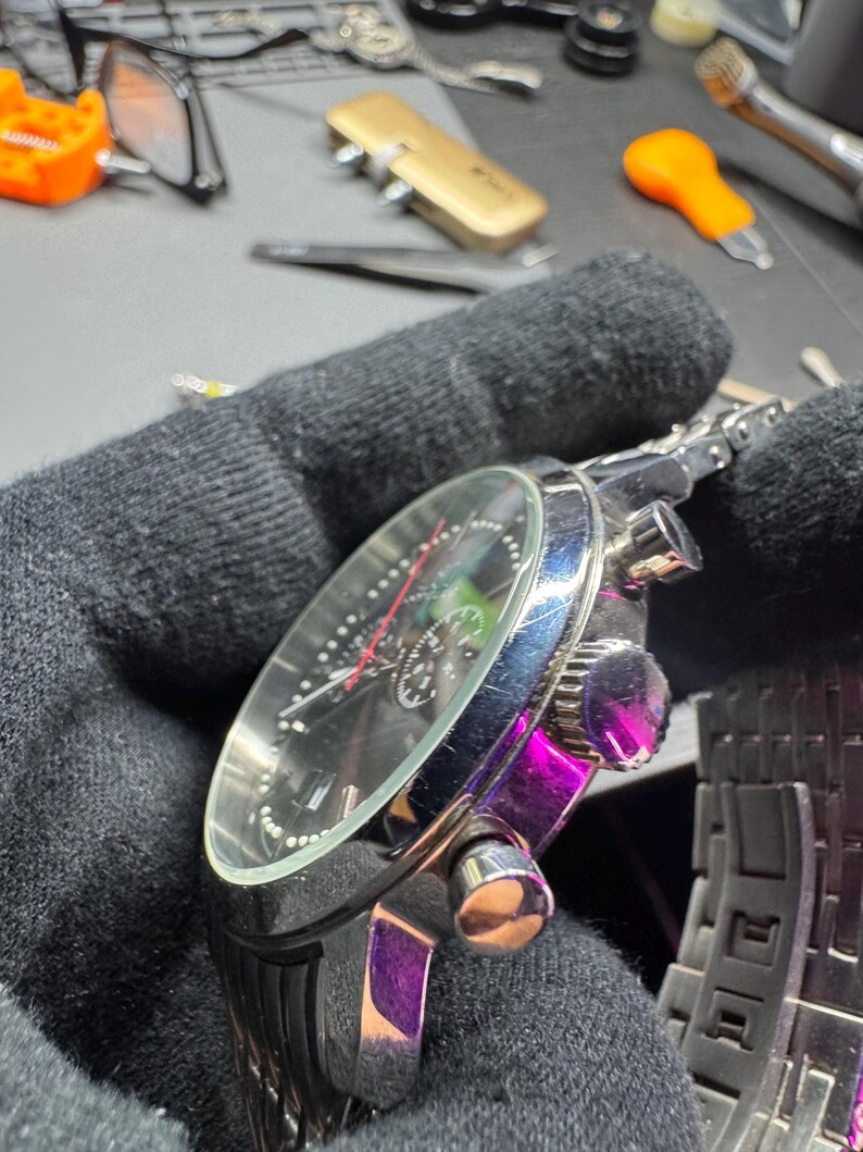 May include: A close-up of a silver and black wristwatch with a black face, featuring silver hands and markers. The watch has a stainless steel band and multiple dials. The watch is held in a black gloved hand.