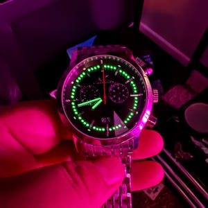 May include: A stainless steel wristwatch with a black face and luminous green accents. The watch features a date display and a chronograph function with red and green hands. The watch is held by a hand.