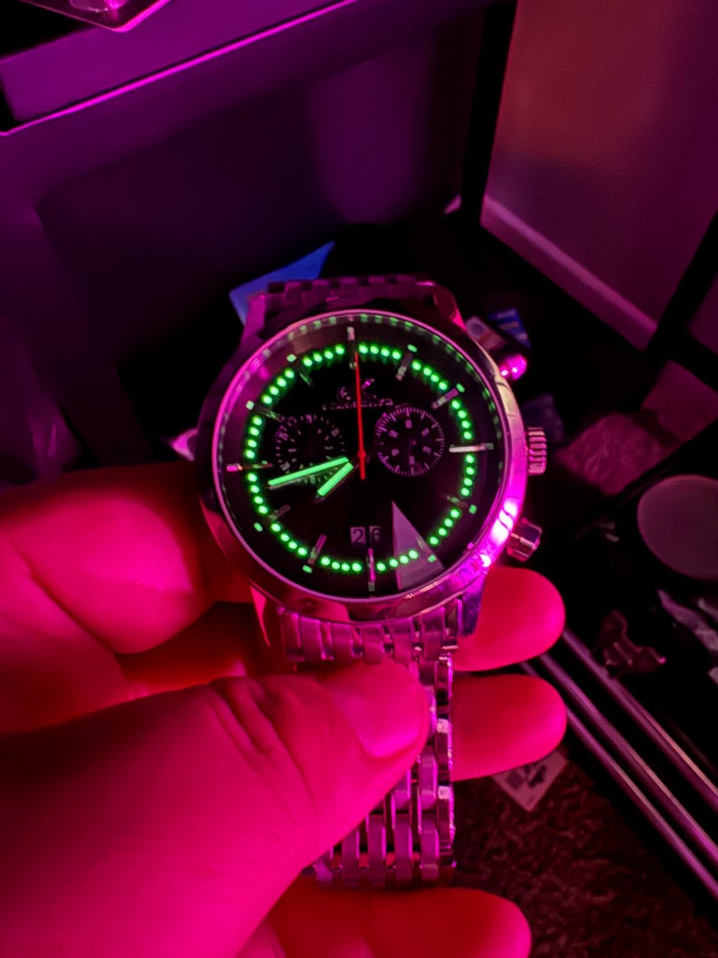 May include: A stainless steel wristwatch with a black face and luminous green accents. The watch features a date display and a chronograph function with red and green hands. The watch is held by a hand.