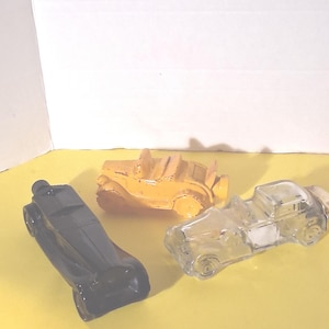 May include: Three vintage car-shaped glass bottles. One is black, resembling a classic car. Another is yellow, a convertible. The third is clear, a pickup truck. All are displayed on a yellow surface.