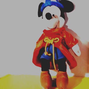 Puede incluir: Un peluche de Mickey Mouse vestido de hechicero, con un sombrero de mago azul con una luna y estrellas, una capa roja, pantalones cortos azules y botas marrones. El juguete est&aacute; sobre una superficie amarilla.