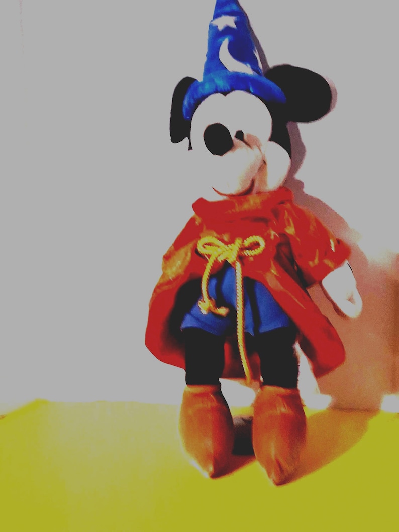 Puede incluir: Un peluche de Mickey Mouse vestido de hechicero, con un sombrero de mago azul con una luna y estrellas, una capa roja, pantalones cortos azules y botas marrones. El juguete est&aacute; sobre una superficie amarilla.