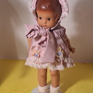 May include: A vintage doll wearing a pink dress, bonnet, and shoes. The dress features a ruffled skirt, lace trim, and floral accents. The doll has brown hair and is standing on a white circular base. The doll's outfit is a soft pink color.