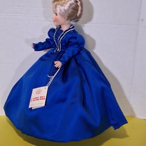May include: A Royal Doll wearing a bright blue satin dress. The doll has blonde hair in an updo and a dress with long sleeves and white trim. The doll is standing on a yellow surface.