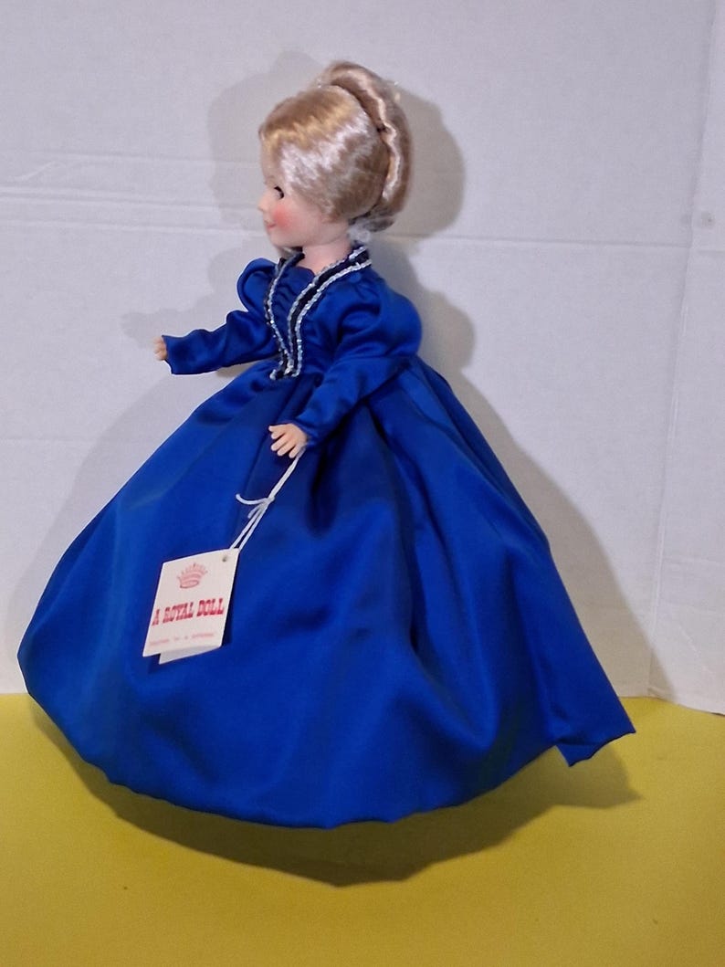 May include: A Royal Doll wearing a bright blue satin dress. The doll has blonde hair in an updo and a dress with long sleeves and white trim. The doll is standing on a yellow surface.