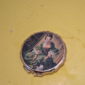 May include: A vintage, round compact mirror with a gold-toned metal frame. The mirror's face features a painted image of a woman in a green dress. The image is set against a light background. The compact is on a yellow surface.