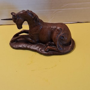 May include: A brown, detailed sculpture of a reclining unicorn with a long, spiraled horn. The mythical creature rests on a textured base, with its head lowered and legs tucked beneath its body. The sculpture is set against a yellow background.