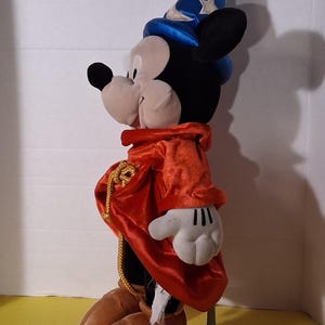 Puede incluir: Un peluche de Mickey Mouse vestido de hechicero, con un sombrero azul con estrellas plateadas, una t&uacute;nica de terciopelo rojo y zapatos marrones. La figura tiene orejas negras, una cara blanca y guantes blancos. Una cuerda dorada adorna la t&uacute;nica.