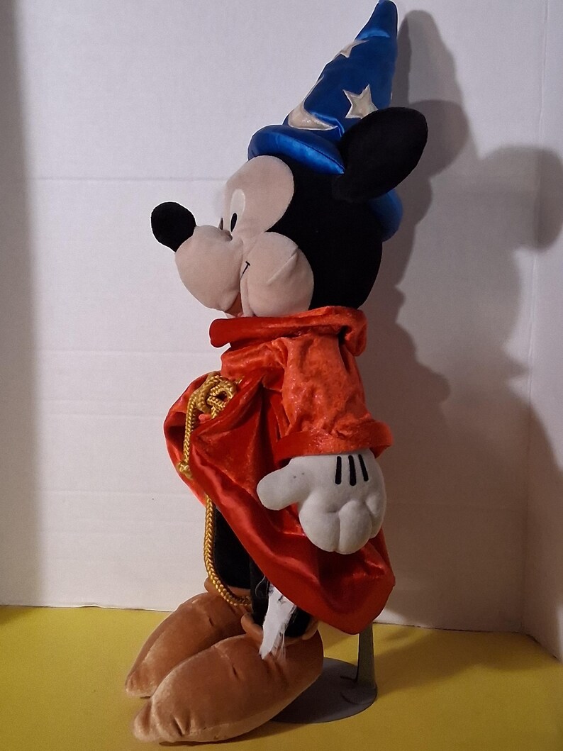 Puede incluir: Un peluche de Mickey Mouse vestido de hechicero, con un sombrero azul con estrellas plateadas, una t&uacute;nica de terciopelo rojo y zapatos marrones. La figura tiene orejas negras, una cara blanca y guantes blancos. Una cuerda dorada adorna la t&uacute;nica.