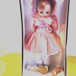 May include: A vintage doll in a box, wearing a red and white striped dress and bonnet. The doll has a pale complexion and is missing a leg. The box is white with a clear front, and the doll is labeled "Japan".
