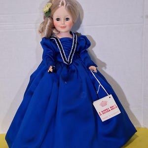 May include: A vintage doll in a blue dress with blonde hair. The doll has a tag that reads "A Royal Doll". The dress features long sleeves and a decorative neckline. A yellow flower is placed in the doll's hair.