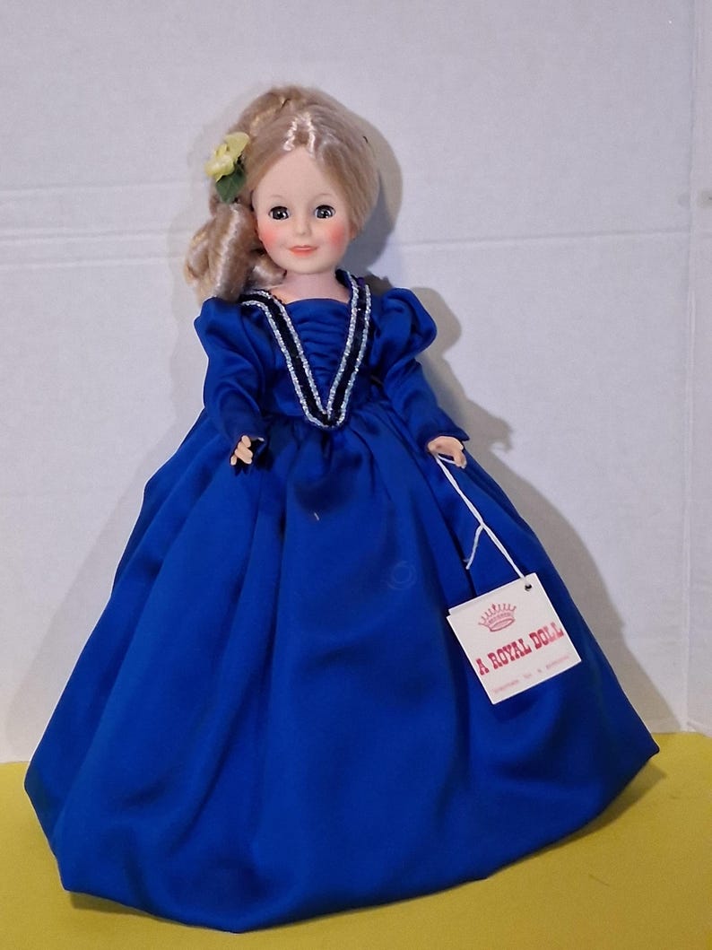 May include: A vintage doll in a blue dress with blonde hair. The doll has a tag that reads "A Royal Doll". The dress features long sleeves and a decorative neckline. A yellow flower is placed in the doll's hair.
