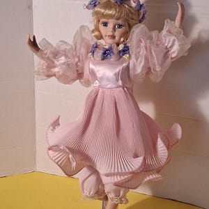 May include: A porcelain doll in a pink pleated dress and matching bloomers, adorned with purple flower accents. The doll has blonde hair and is posed with arms outstretched, wearing ballet slippers. The doll is on a silver stand.