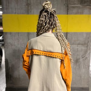 Könnte beinhalten: Beiger Blazer mit orangefarbenen Ärmeln und Ziernähten. Auf der Rückseite befindet sich ein orangefarbenes Panel mit braunen Nähten. Der Text "OPEN UP" ist auf einem schwarzen Patch unten am Blazer zu sehen.
