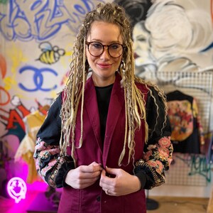 Può includere: Un gilet bordeaux con chiusura a bottoni, indossato sopra un top nero con maniche ricamate floreali. Lo sfondo presenta graffiti e una faccina sorridente al neon.