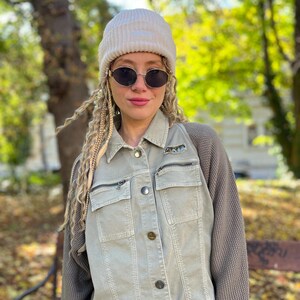 May include: A woman wearing a light green jacket with a knit sweater attached to the sleeves, a cream-coloured beanie, and oval sunglasses. The jacket has two zipped pockets and button closures. The background features a park setting with trees.