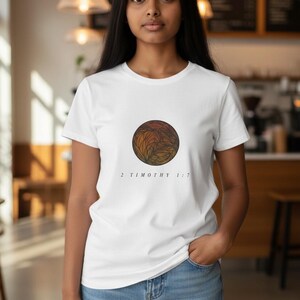 May include: White t-shirt with a circular design of orange, brown, and yellow leaf patterns. The text "2 Timothy 1:7" is printed below. The person is wearing blue jeans.