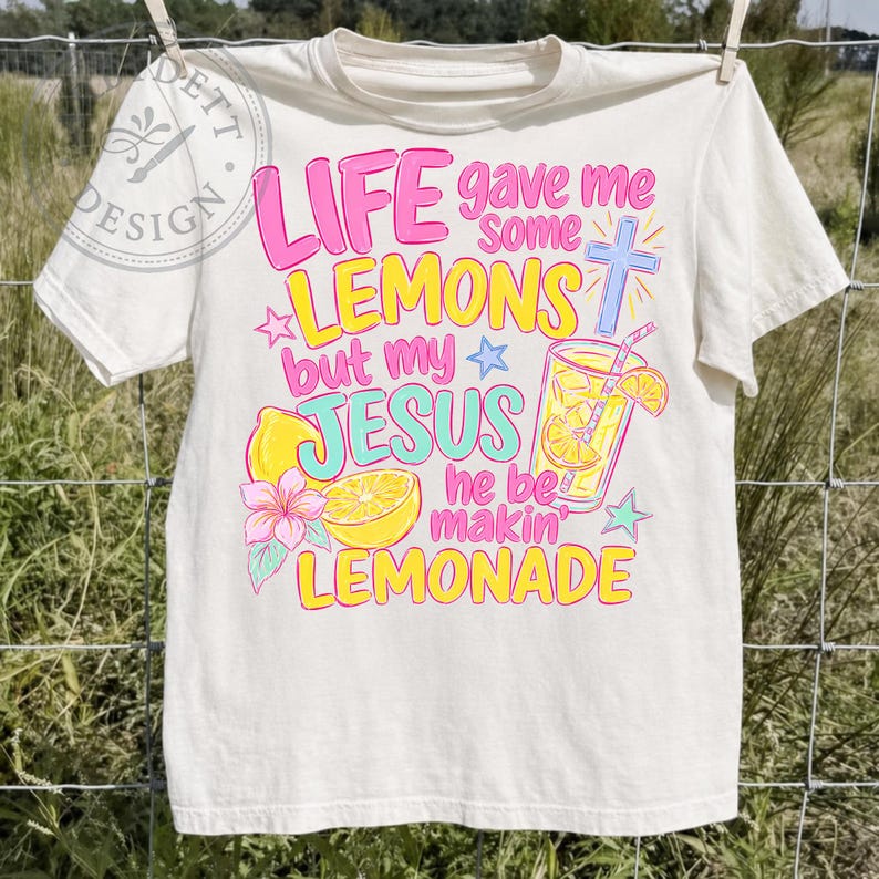 May include: A cream-colored short-sleeve t-shirt featuring a colorful graphic with the text "LIFE gave me some LEMONS but my JESUS he be makin' LEMONADE". The design includes illustrations of lemons, a glass of lemonade, a cross, and stars.