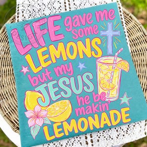 May include: A teal t-shirt featuring the text LIFE gave me some LEMONS but my JESUS he be makin' LEMONADE in pink and yellow lettering. The design includes a light blue cross, a glass of lemonade with a straw, two yellow lemons, and a pink flower.