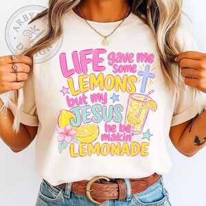 May include: A cream-colored t-shirt featuring a colorful graphic with the text "LIFE gave me some LEMONS but my JESUS he be makin' LEMONADE". The design includes illustrations of yellow lemons, a glass of lemonade, a blue cross, and pink flowers.