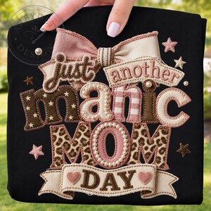 以下が含まれることがあります： 「just another manic MOM DAY」という文字が刺繍された、質感のあるアップリケデザインの黒いTシャツ。大きなリボン、星、ハートが茶色、ピンク、クリーム色で描かれ、フェイクパールとヒョウ柄のアクセントが施されています。