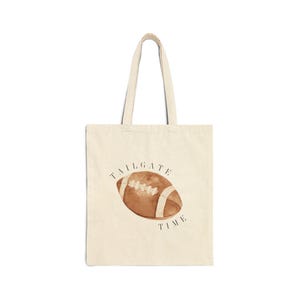 May include: A cream-colored canvas tote bag featuring a watercolor illustration of a brown American football centered on the front. The text TAILGATE TIME is printed in a simple black serif font, arching over and under the football graphic.