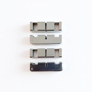 May include: Four silver-colored metal guitar nut pieces arranged vertically on a white background. Each piece has a rectangular shape with a series of square cutouts. The bottom two pieces have an "R" etched on them.