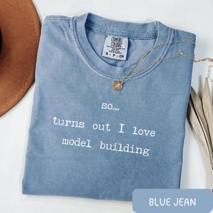 May include: A blue jean-colored t-shirt with the text "so... turns out I love model building" in white. The shirt has a crew neck and short sleeves. A gold necklace with a pendant is draped across the shirt.