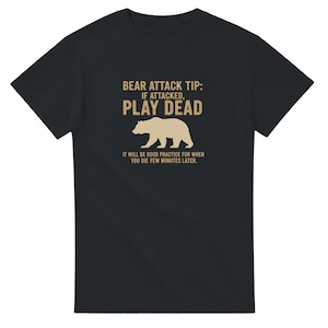 Peut inclure: T-shirt noir avec un graphisme humoristique. Le motif présente le texte "BEAR ATTACK TIP: IF ATTACKED, PLAY DEAD" au-dessus d'une silhouette d'ours. En dessous, on peut lire "IT WILL BE GOOD PRACTICE FOR WHEN YOU DIE FEW MINUTES LATER."