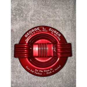 May include: A round, red ashtray with the text "GEORGE L. ZUBER" and "For The Finest In Corn Fed Turkeys". The ashtray has a ridged design and is made of a shiny, metallic material. The text includes a phone number and address.