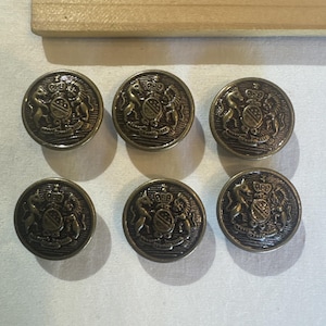 May include: A set of six round, metallic buttons featuring an embossed heraldic design. Each button displays a central shield flanked by two rampant animals, topped with a crown, and finished in a dark, antique bronze-toned metal.