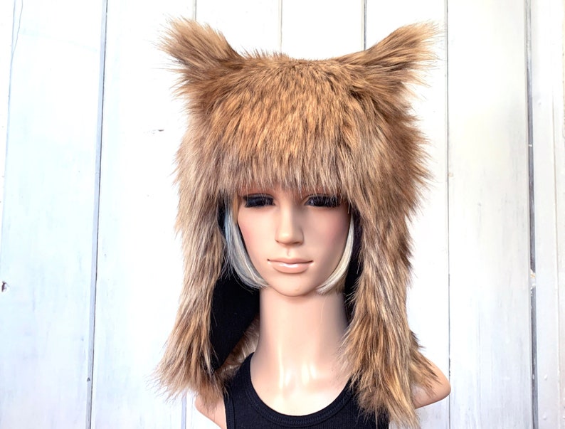 May include: A brown faux fur hat with long ear flaps and a shaggy texture. The hat is worn on a mannequin head.