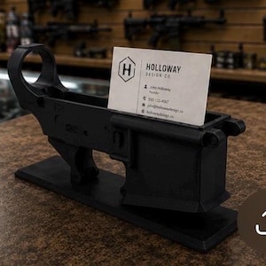 May include: A matte black AR-15 lower receiver shaped business card holder mounted on a rectangular base. A white business card is inserted into the magazine well, featuring the text HOLLOWAY DESIGN CO. and a logo with the letter H inside a hexagon.