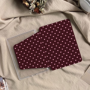 以下が含まれることがあります： ピンクのハート模様のボルドー色のタブレットケース。ケースは開いており、透明なプラスチック製のタブレットホルダーが現れています。デザインはカバーとケースの裏側に繰り返されています。ケースはベージュ色の表面にあります。