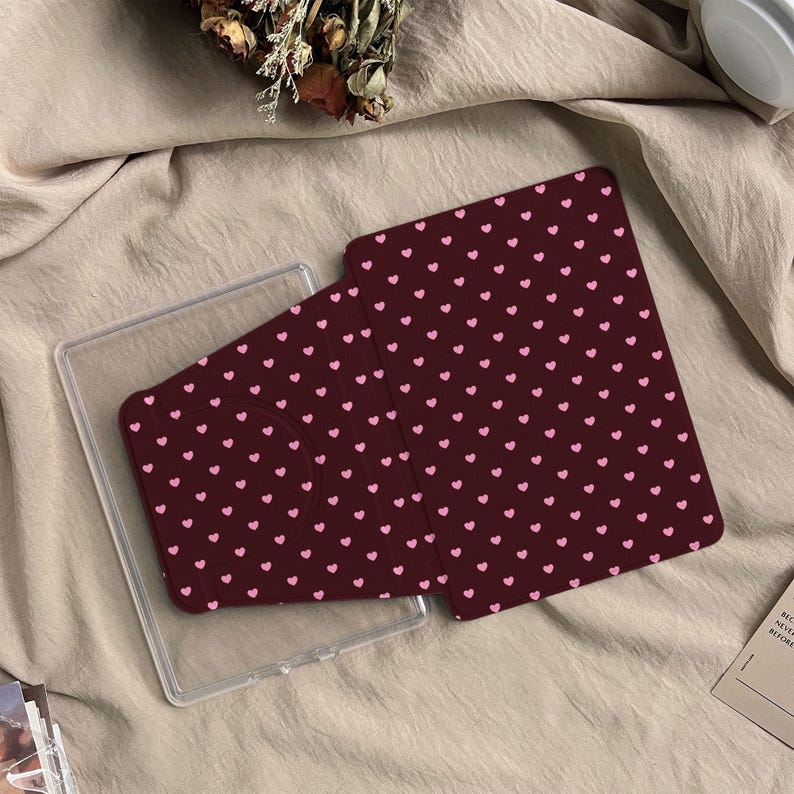 May include: A dark maroon tablet case featuring a repeating pattern of small light pink hearts. The case has a rectangular shape with a circular cutout on the left side and is positioned partially inside a clear plastic protective shell.