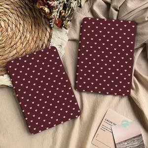 May include: Two rectangular e-reader cases featuring a dark maroon background with a repeating pattern of small light pink hearts. The cases are positioned on a textured fabric surface next to a small card with the text GROWTH.