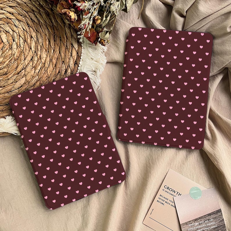 May include: Two rectangular e-reader cases featuring a dark maroon background with a repeating pattern of small light pink hearts. The cases are positioned on a textured fabric surface next to a small card with the text GROWTH.